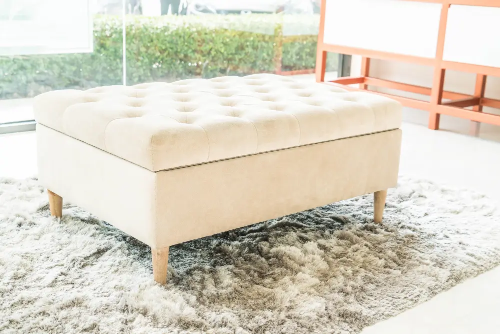 A white ottoman placed on a rug, positioned in front of a window allowing natural light to enter the room.